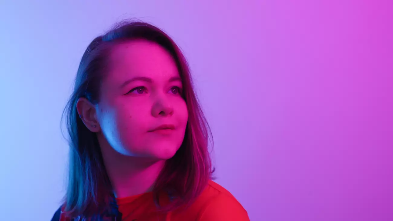 The photo shows the composer Oktawia Paczkowska looking slightly upward with a calm expression. The background is softly lit in a gradient of blue on one side and pink-purple on the other, creating a vibrant and colorful atmosphere.