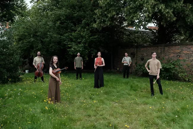 six musicians of Ear to the Earth ensemble standing in a green field. A violinist, pianist, percussionist, cellist, flutist, and clarinetist stand spaced out across the green grass. A backdrop of a garden wall, a pair of trees, and flowers accent the background