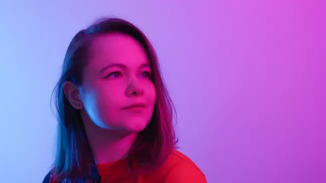 The photo shows the composer Oktawia Paczkowska looking slightly upward with a calm expression. The background is softly lit in a gradient of blue on one side and pink-purple on the other, creating a vibrant and colorful atmosphere.